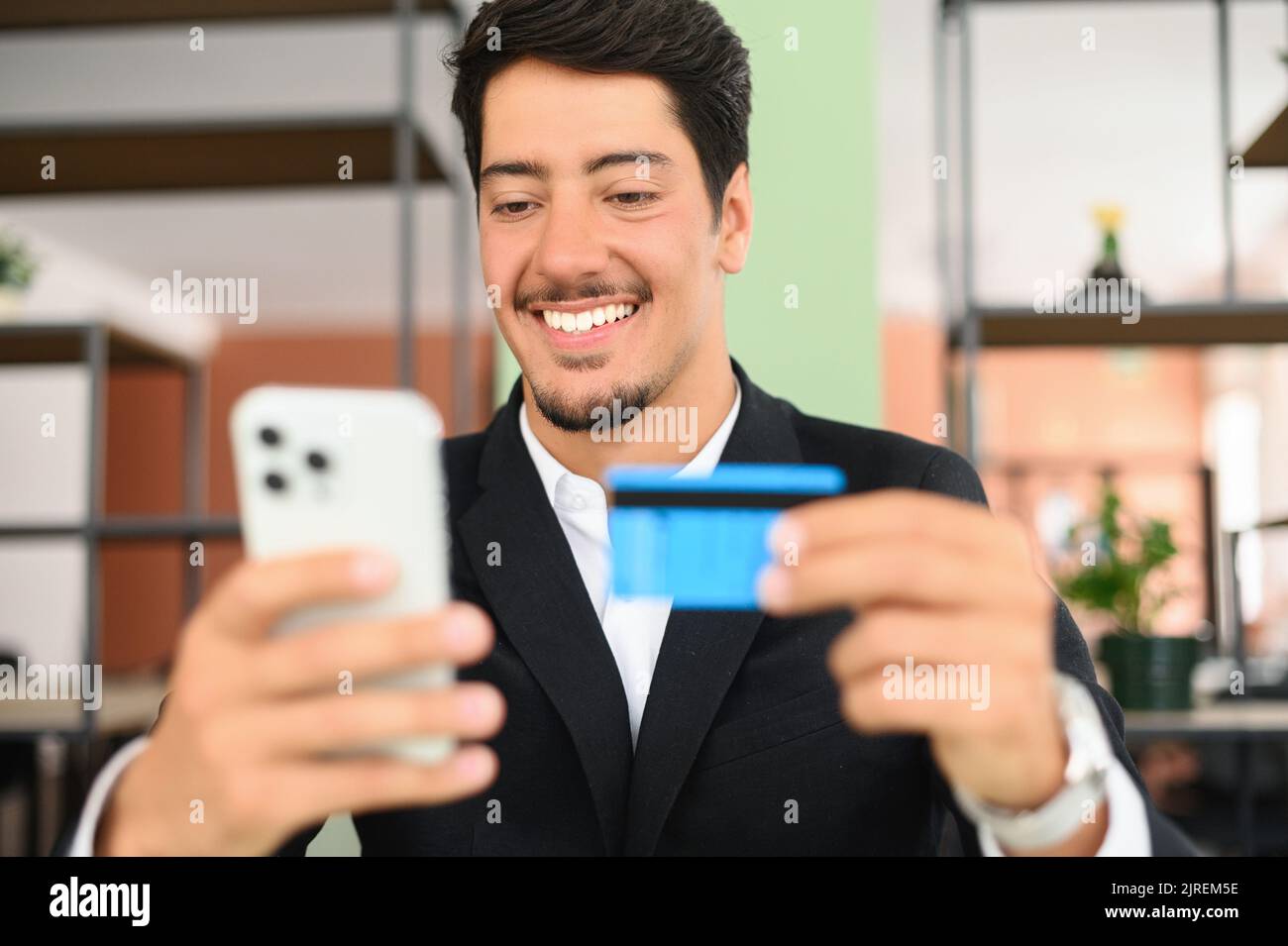 Fröhlicher lateinischer Mann macht online kaufen. Hispanic Kerl in Business Wear hält Telefon und eine Kreditkarte, macht Geldtransaktion online sitzen im Büro, Kerl ist mit Smartphone und Debit-Bankkarte Stockfoto