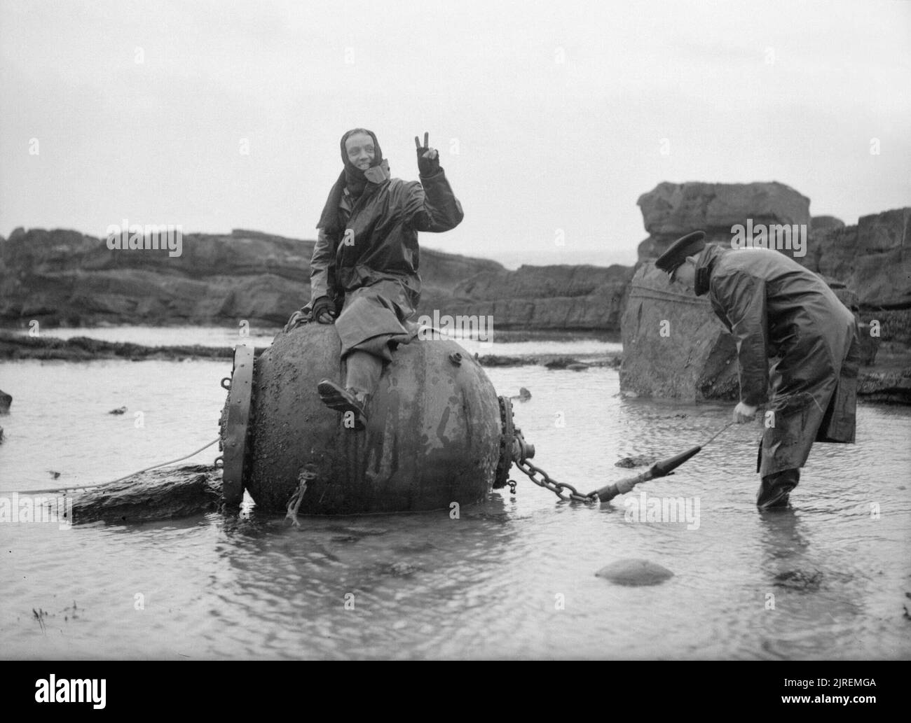 Die Royal Navy während des Zweiten Weltkriegs bekleidet mit Wolle Helme, Öl Skins, und Meer Stiefel, die meine Bewertung, Mitglied einer Marine mine Entsorgung Partei das V-Zeichen auf eine Mine sitzen, die als ein weiterer Tage Arbeit in Anstruther, Schottland getan ist. Stockfoto