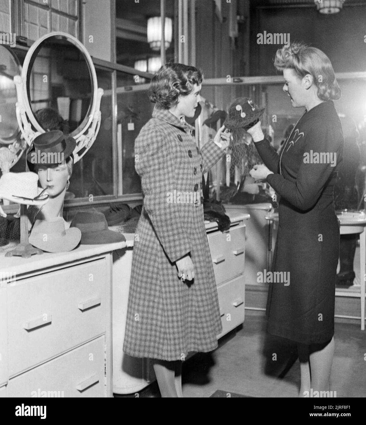 Hilda Chilingworth (links) schaut auf Hüte in einem Geschäft während Ihrer Mittagspause Brea in London während 1942. Hilda Chilingworth (links) wird durch einen Assistenten unterstützt, als sie Hüte in einem Geschäft während Ihrer Mittagspause aussieht. Obwohl Sie die Speicherung hat einen Hut zu kaufen, die Preise sind zu hoch, und Sie schließlich verlässt den Laden leer aus. Stockfoto