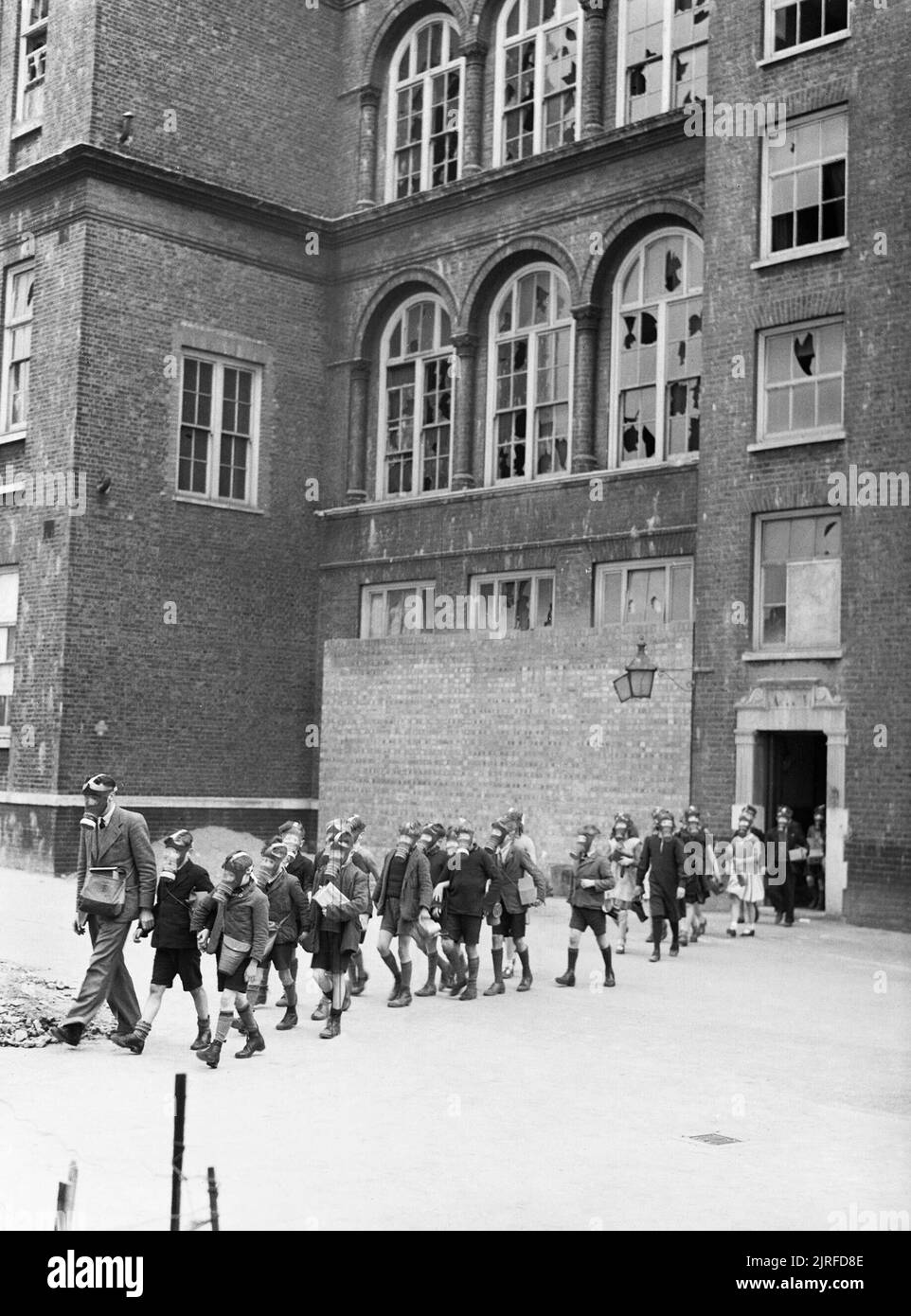 Anti-gas Bohren an der Alten Woolwich Road School in Greenwich, London 1941. Eine Lehrerin einer Gasmaske führt eine große Gruppe von Kindern aus dem Eingang der alten Woolwich Road School in Greenwich, London. Die Kinder werden über den Spielplatz in einer Zeile zwei Arches und alle tragen ihre Gasmasken. Die Schule hat deutlich beschädigt wurde durch Luftangriffe, wie viele Fenster haben Scheiben fehlt oder zertrümmert. Es gibt auch einige Schäden am Mauerwerk. Stockfoto