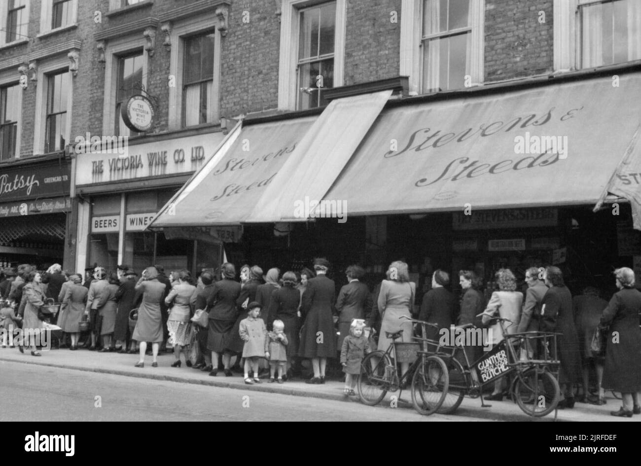 Frauen und Kinder Warteschlange Gemüse von einem Obst- und Gemüsehändler in London während 1945 zu kaufen. Frauen und Kinder Warteschlange Vergangenheit mehrere Geschäfte waren von "Kaufen Patsy" der Gemüsehändler, auf einer Einkaufsstraße, irgendwo in London. Deutlich sichtbar sind die "Victoria Wein Co Ltd.' und 'Stevens und Ross'. Außerhalb des Letzteren, in der rechten unteren Ecke des Fotos, sind zwei Fahrräder mit einem Schild mit der Aufschrift "Gunther lustige gute Metzger". Stockfoto