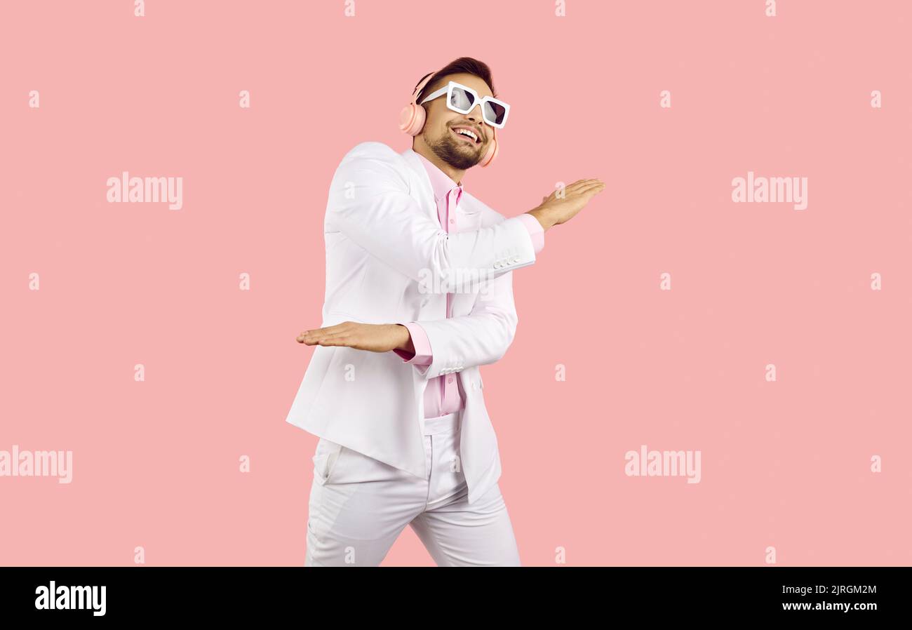 Fröhlicher, lustiger junger Mann in einem weißen Anzug, einem rosa Hemd und einer Sonnenbrille mit kabellosen pinken Kopfhörern hört Musik und tanzt in einem pinken Studio Stockfoto