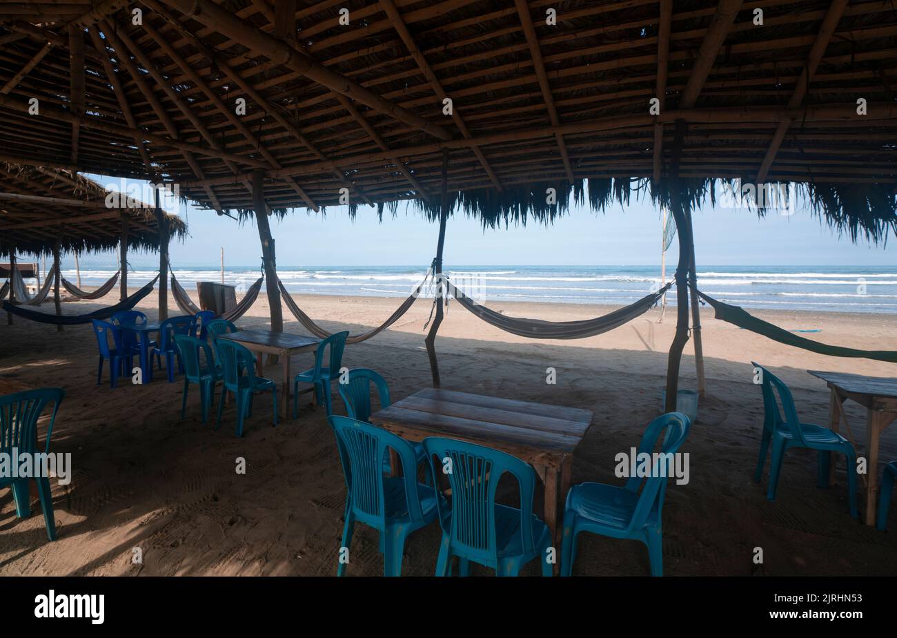 Blick auf ein rustikales Restaurant ohne Menschen, mit einem Bambusdach, Holztischen, Plastikstühlen und Hängematten, die an Stangen am Strand an einem sonnigen Ort hängen Stockfoto