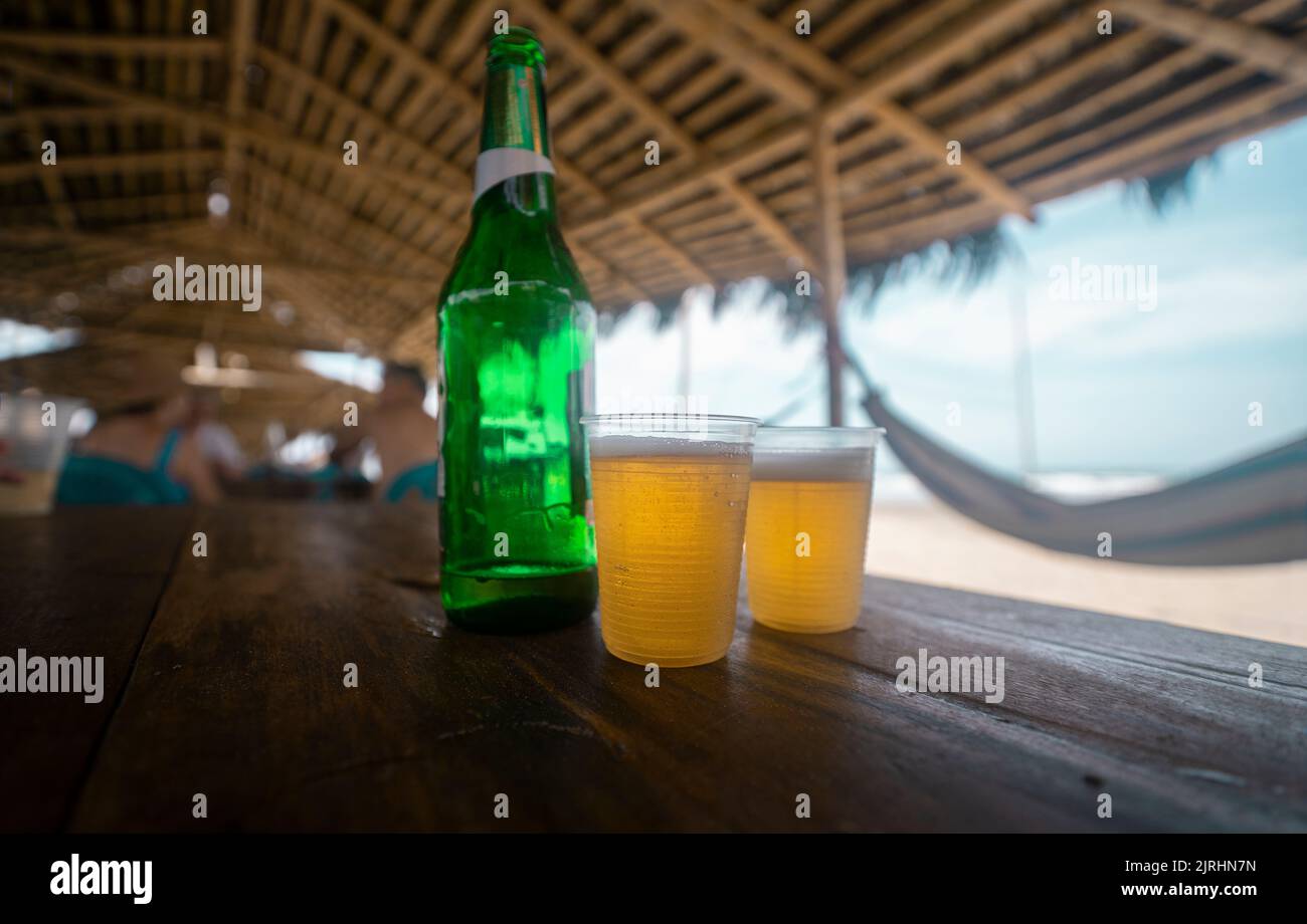 Zwei Plastikgläser mit Bier und einer grünen Flasche auf einem Holztisch in einem Strandrestaurant mit Bambusdach und Hängematten an den Seiten während eines sonnigen D Stockfoto