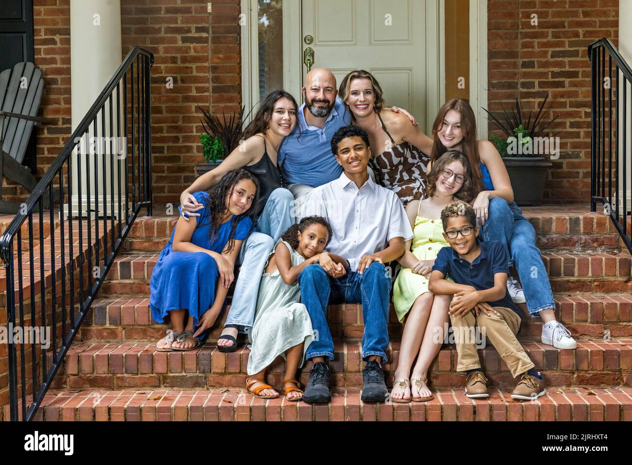 Eine große, gemischte und glückliche Familie, die auf den vorderen Stufen ihres Backsteinhauses in den Vororten sitzt Stockfoto