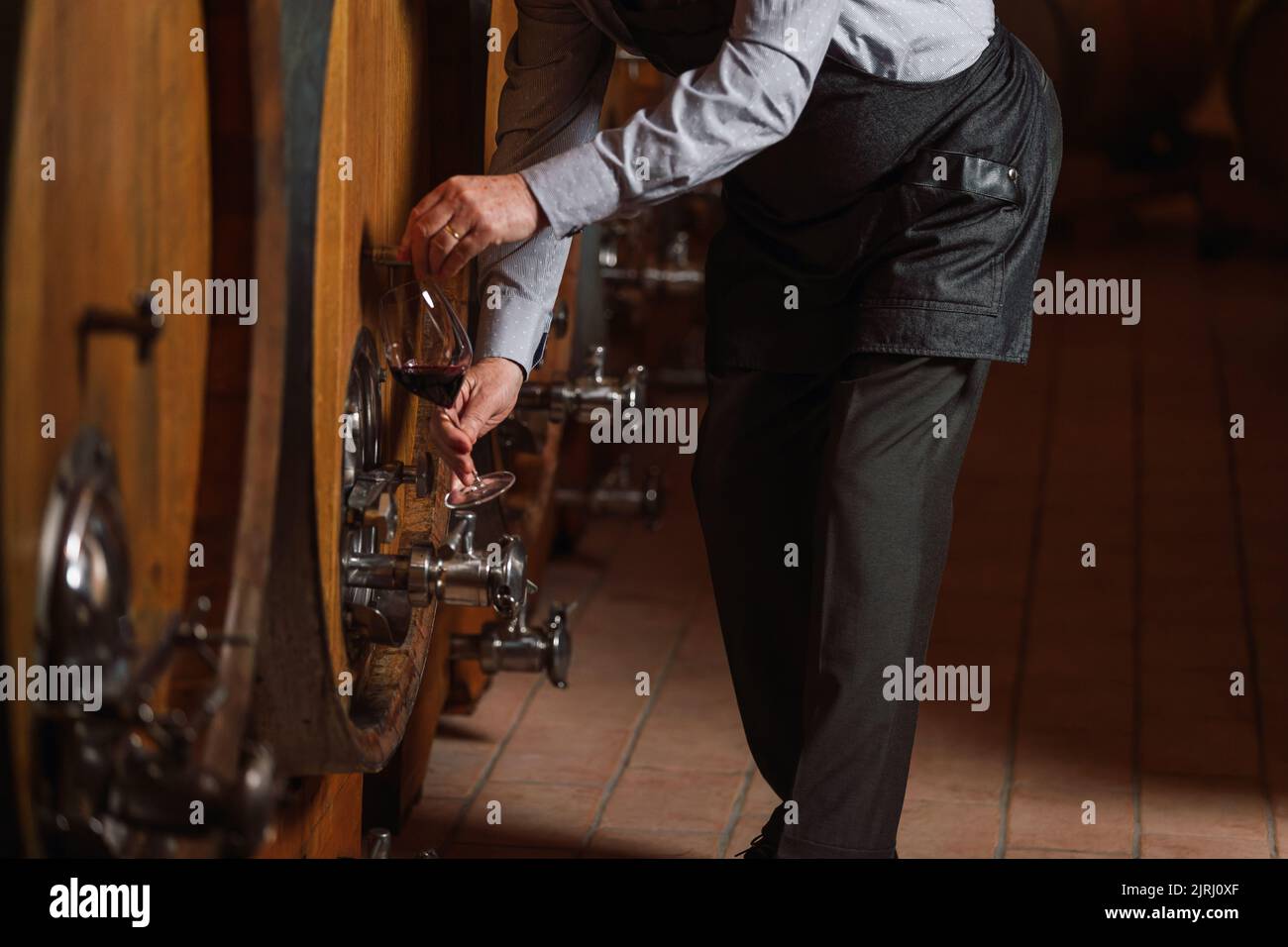 Kaukasischer Senior-Graubart-Winzer, der Rotwein verkostet, um seine Qualität und seinen Geschmack zu ermitteln, steht in der Nähe eines großen Eichenfasses Stockfoto