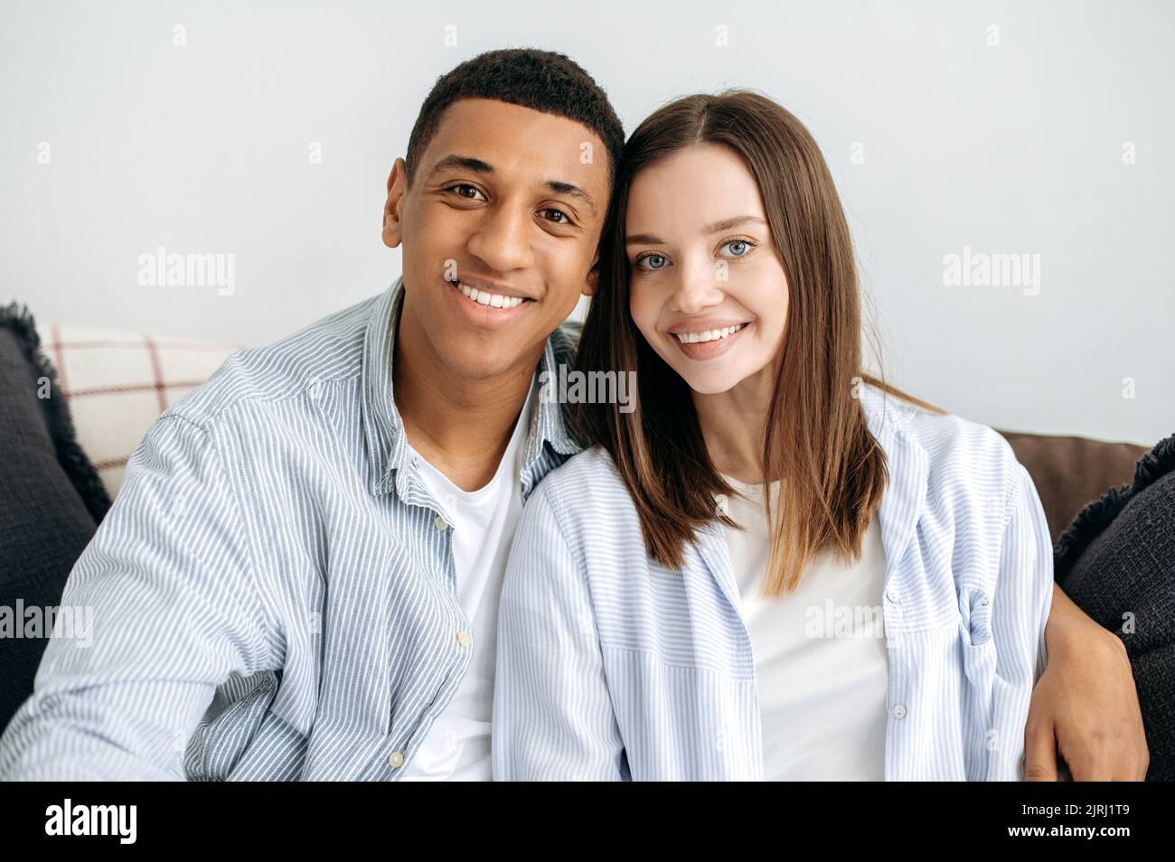 Porträt eines glücklichen verheirateten jungen Paares. Ein positives, multirassisches Paar in Liebe, eine kaukasische Frau und ein hispanischer Mann, die zu Hause im Wohnzimmer auf dem Sofa sitzen, umarmen, die Kamera betrachten und lächeln Stockfoto