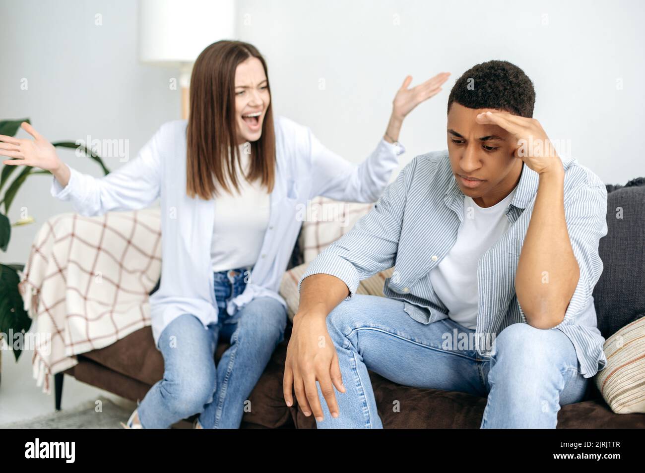 Missverständnisse in Beziehungen, Eifersucht, Streit. Ein verheiratetes, multirassisches Paar sortiert Dinge aus, während eine kaukasische Frau im Wohnzimmer auf einem Sofa sitzt und ihren hispanischen Ehemann anschreit Stockfoto