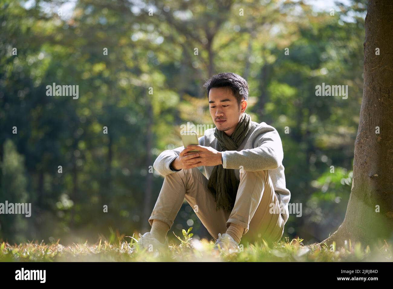 Junger asiatischer Erwachsener, der im Park auf Gras sitzt und auf das Mobiltelefon schaut Stockfoto