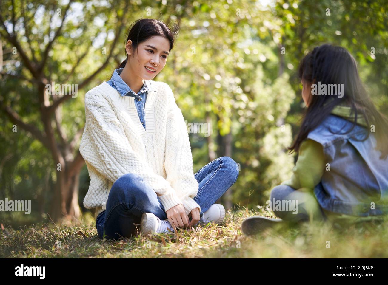 Junge asiatische Mutter sitzt auf Gras im Park mit einem angenehmen Gespräch mit Vorschultochter Stockfoto