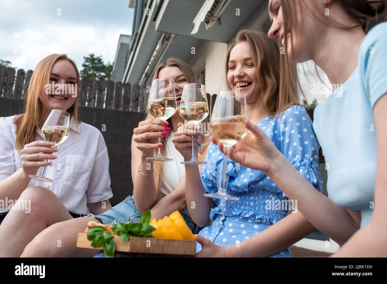 Portrait von jungen lustigen schönen Frauen, die geschnittenes Gemüse, klirrende Gläser Weißwein halten und in der Nähe des Hauses sitzen. Stockfoto