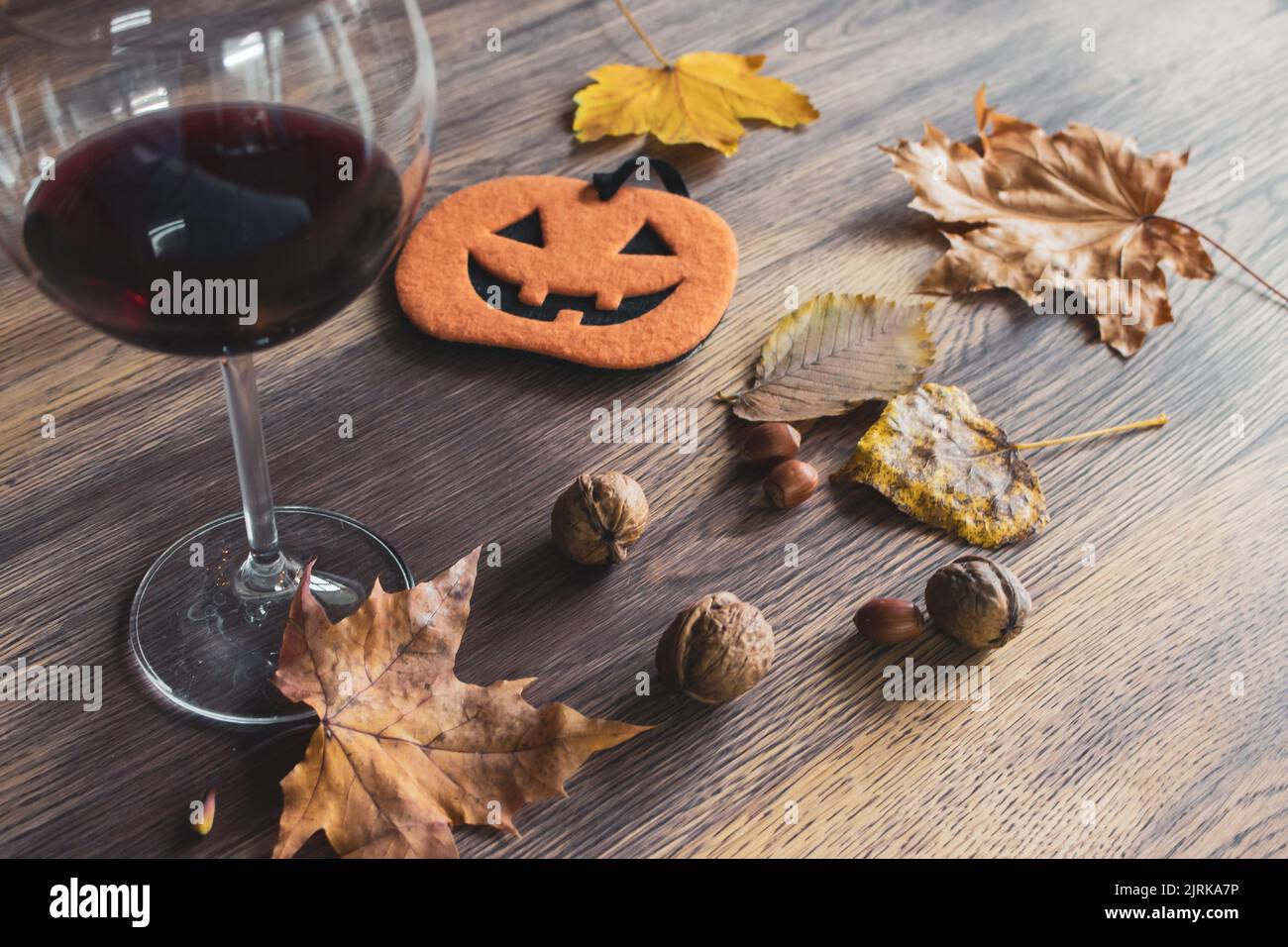 Halloween Stillleben. Ein Glas Rotwein, Nüsse, trockene Blätter und lustige Kürbisfigur auf dem Tisch. Herbstferien. Oktober Hintergrund. Stockfoto