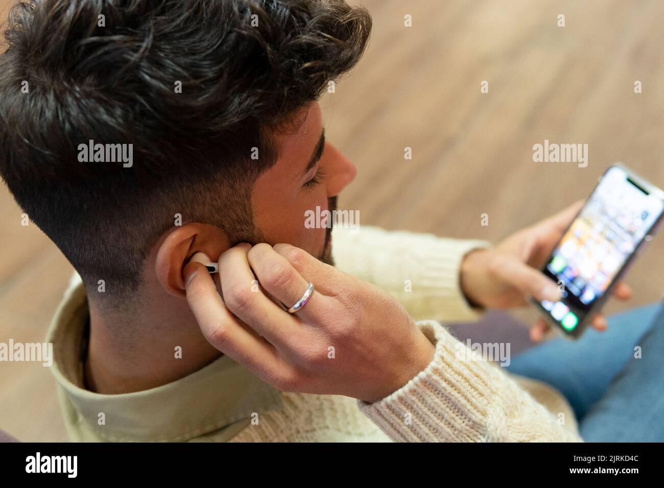 Von oben junger hispanischer Mann mit Bart und dunklem Haar, der zu Hause sitzt und mit kabellosen Kopfhörern auf dem Mobiltelefon surft Stockfoto