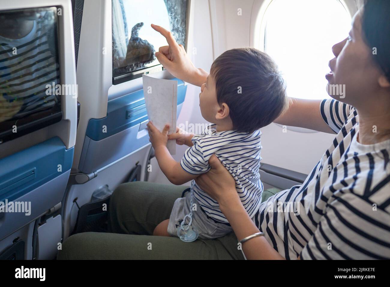 Kleinkind, das im Flugzeug sitzt, auf seinem MutterSchoß und den Bildschirm des Flugunterhaltungssitzes benutzt. Ein Jahr alter Junge, der im Flugzeug fliegt und versucht zu en Stockfoto