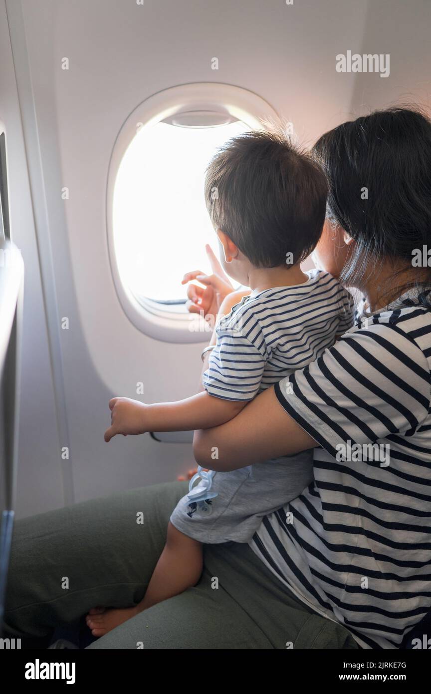 Das Kleinkind, das im Flugzeug sitzt und auf seinem MutterSchoß sitzt, blickt beide aus einem Flugzeugfenster. Ein Jahr alter Junge, der im Flugzeug fliegt und versucht zu fliegen Stockfoto