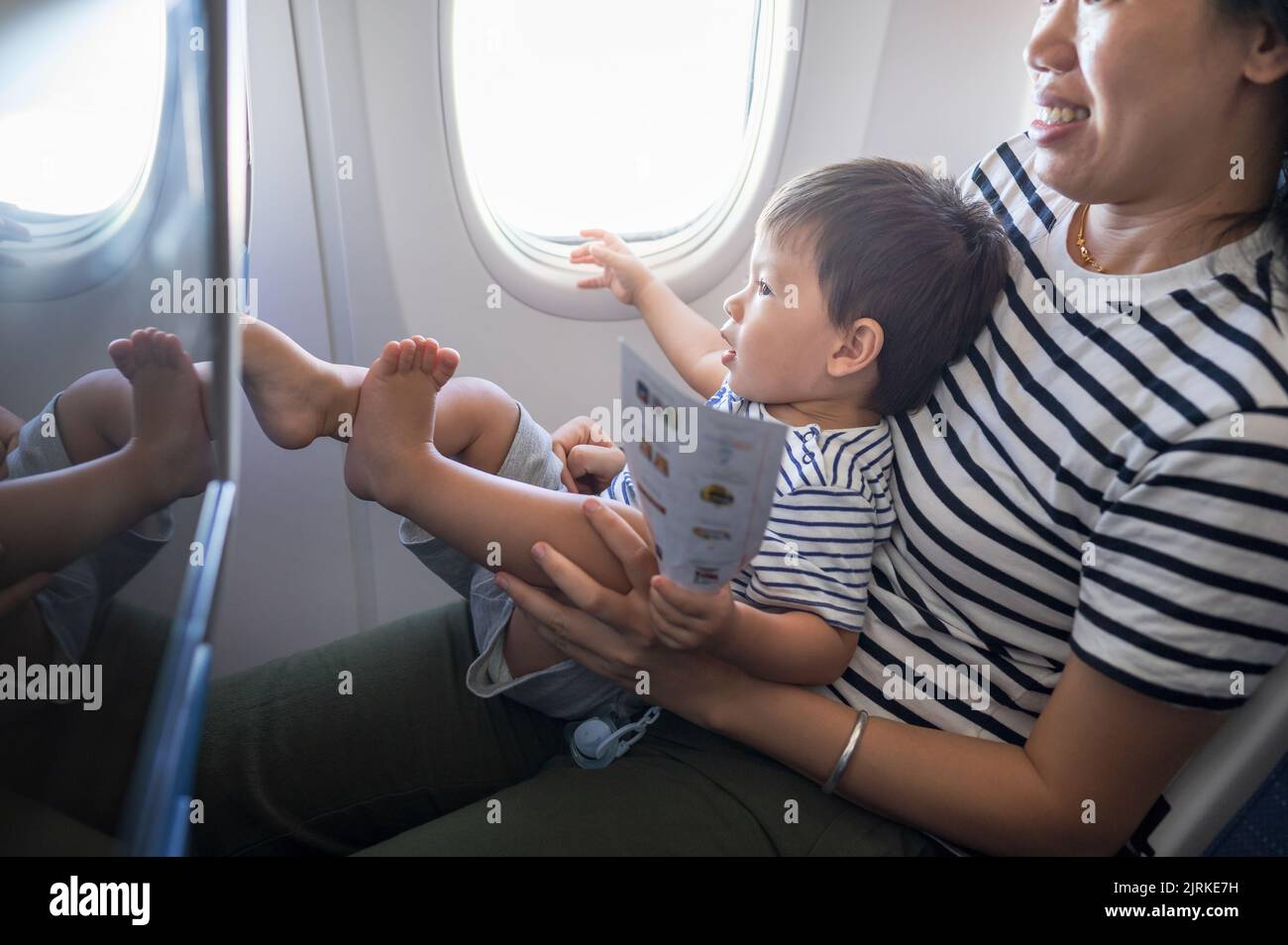 Kleinkind, das im Flugzeug sitzt, auf seinem MutterSchoß und den Bildschirm des Flugunterhaltungssitzes benutzt. Ein Jahr alter Junge, der im Flugzeug fliegt und versucht zu en Stockfoto