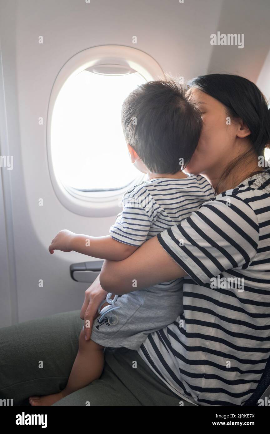 Das Kleinkind, das im Flugzeug sitzt und auf seinem MutterSchoß sitzt, blickt beide aus einem Flugzeugfenster. Ein Jahr alter Junge, der im Flugzeug fliegt und versucht zu fliegen Stockfoto