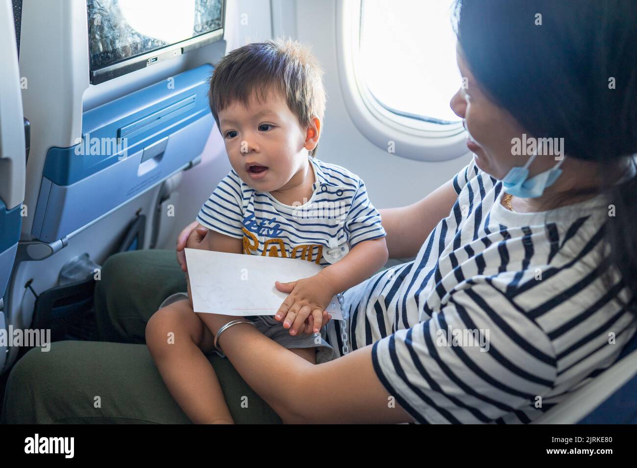 Kleinkind, das im Flugzeug auf dem MutterSchoß sitzt. Ein Jahr alter Junge, der im Flugzeug fliegt und versucht, sich zu unterhalten Stockfoto