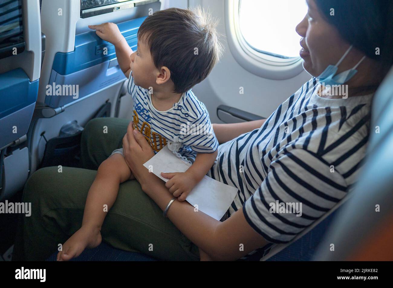 Kleinkind, das im Flugzeug sitzt, auf seinem MutterSchoß und den Bildschirm des Flugunterhaltungssitzes benutzt. Ein Jahr alter Junge, der im Flugzeug fliegt und versucht zu en Stockfoto