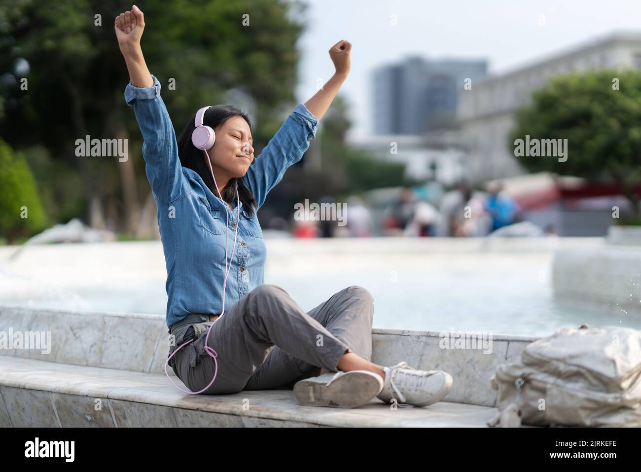 Ein ganzer Körper entspannter Frauen, die mit geschlossenen Augen Musik in Kopfhörern hören und die Fäuste hochheben, während sie in der Nähe des Brunnens im Park sitzen Stockfoto