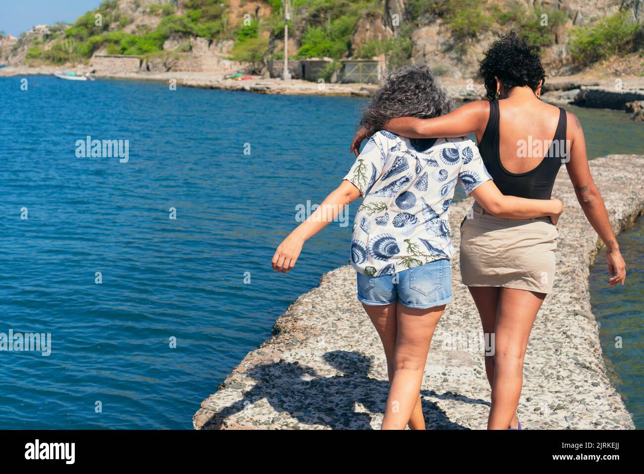 Rückansicht von anonymen besten ethnischen Freundinnen, die sich umarmen, während sie auf dem Steindock im Ozean gegen den Berg gehen Stockfoto