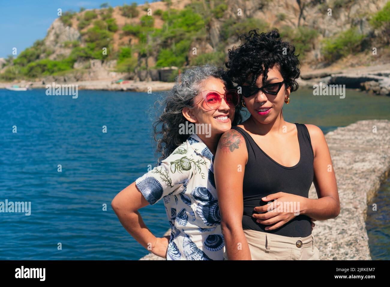 Seitenansicht der besten ethnischen Freundinnen, die sich umarmen, während sie auf dem Steindock im Ozean gegen den Berg stehen und die Kamera betrachten Stockfoto