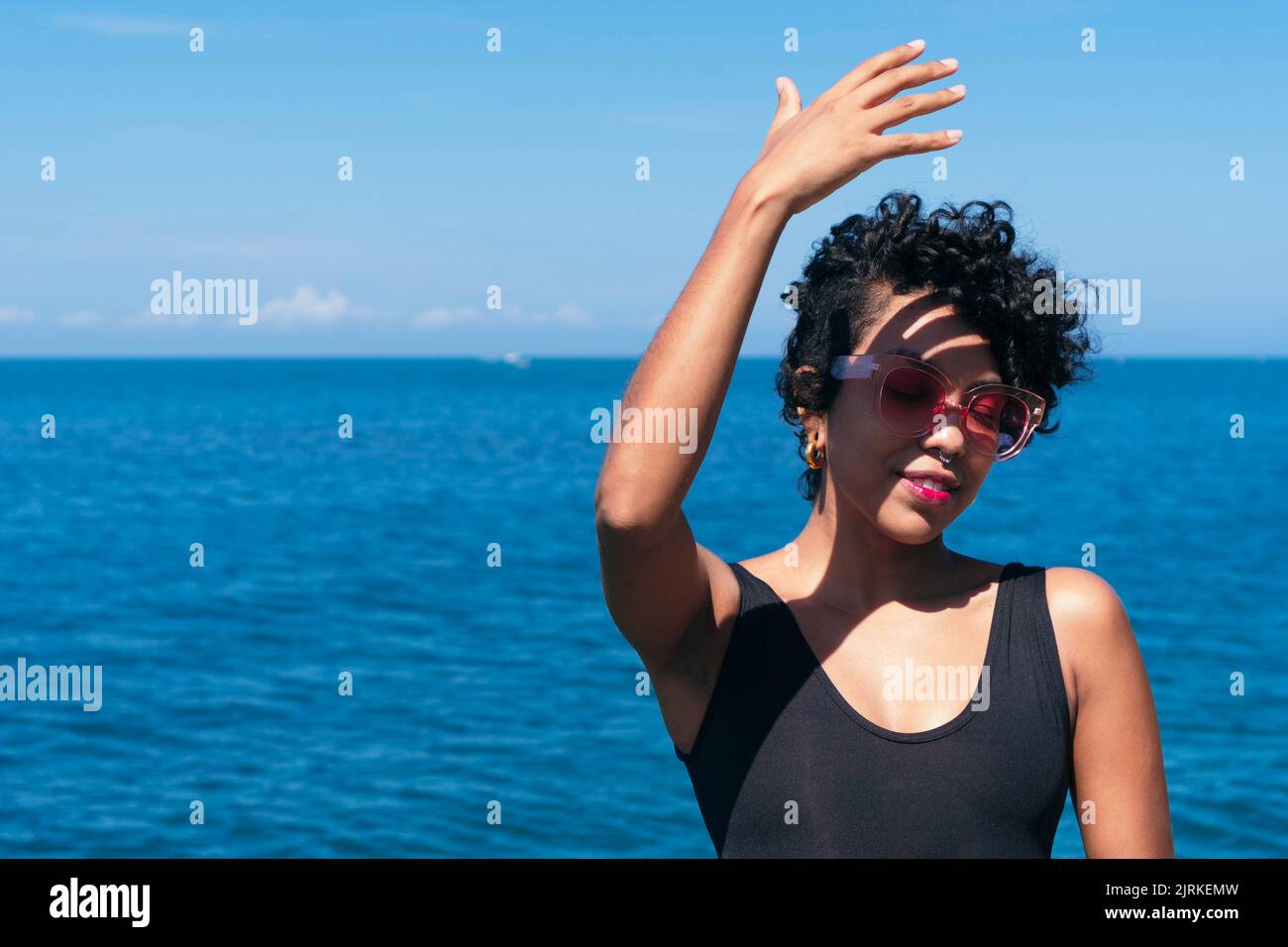 Fröhliche afroamerikanische Frau mit lockigen Haaren, die mit dem Arm nach oben steht, während er unter blauem Himmel einen Schatten auf sein Gesicht gegen den Ozean wirft Stockfoto