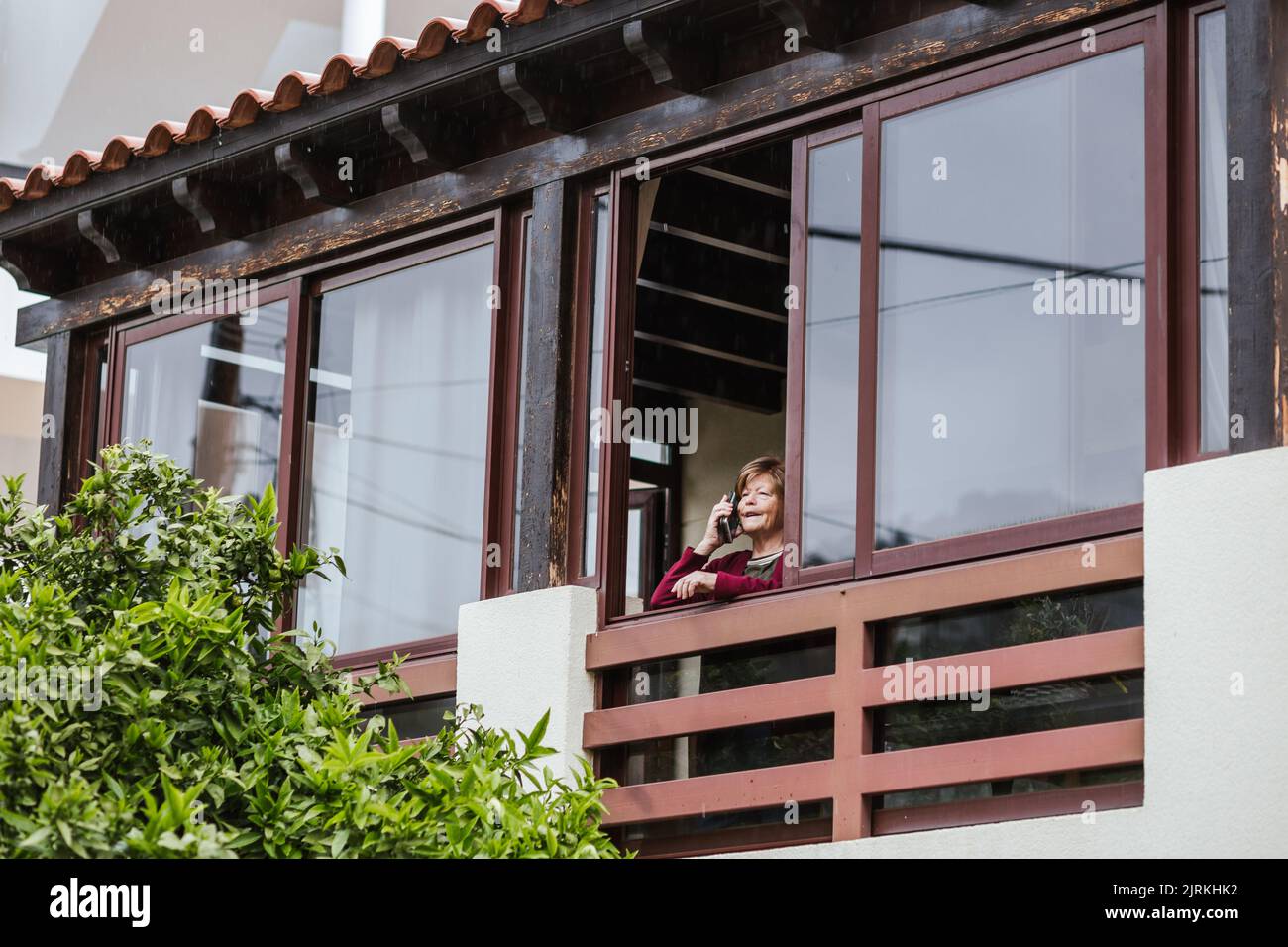 Von unten eine ältere Dame, die zu Hause neben dem Fenster steht und auf dem Balkon einen Anruf entgegennimmt Stockfoto