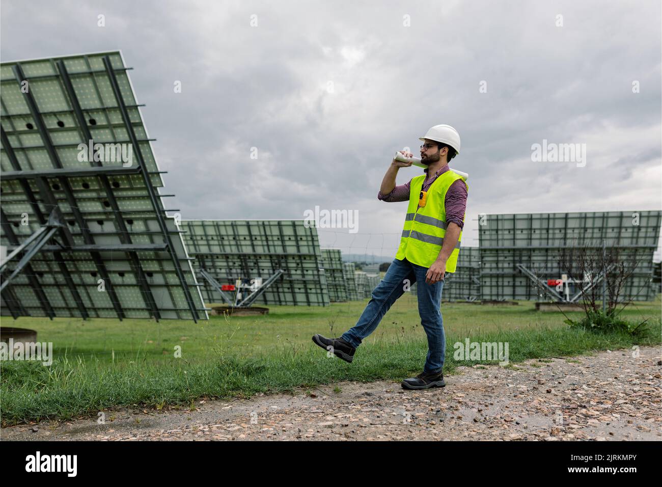 Ganzkörpermensch in Weste und Hardhut, der Zugluft trägt und am bewölkten Tag in der Nähe von Photovoltaikanlagen auf dem Weg geht Stockfoto