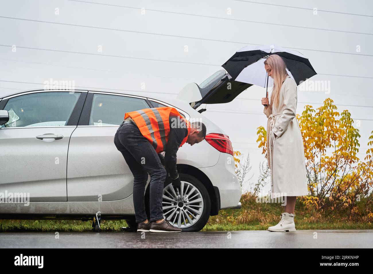 Schöne junge Frau, die neben dem Auto steht und einen Regenschirm hält, während der Mechaniker die Radmuttern abschraubt. Junger Mann in Weste Reparatur Frau Auto auf der Straße. Konzept des Notdienstes im Straßenverkehr. Stockfoto