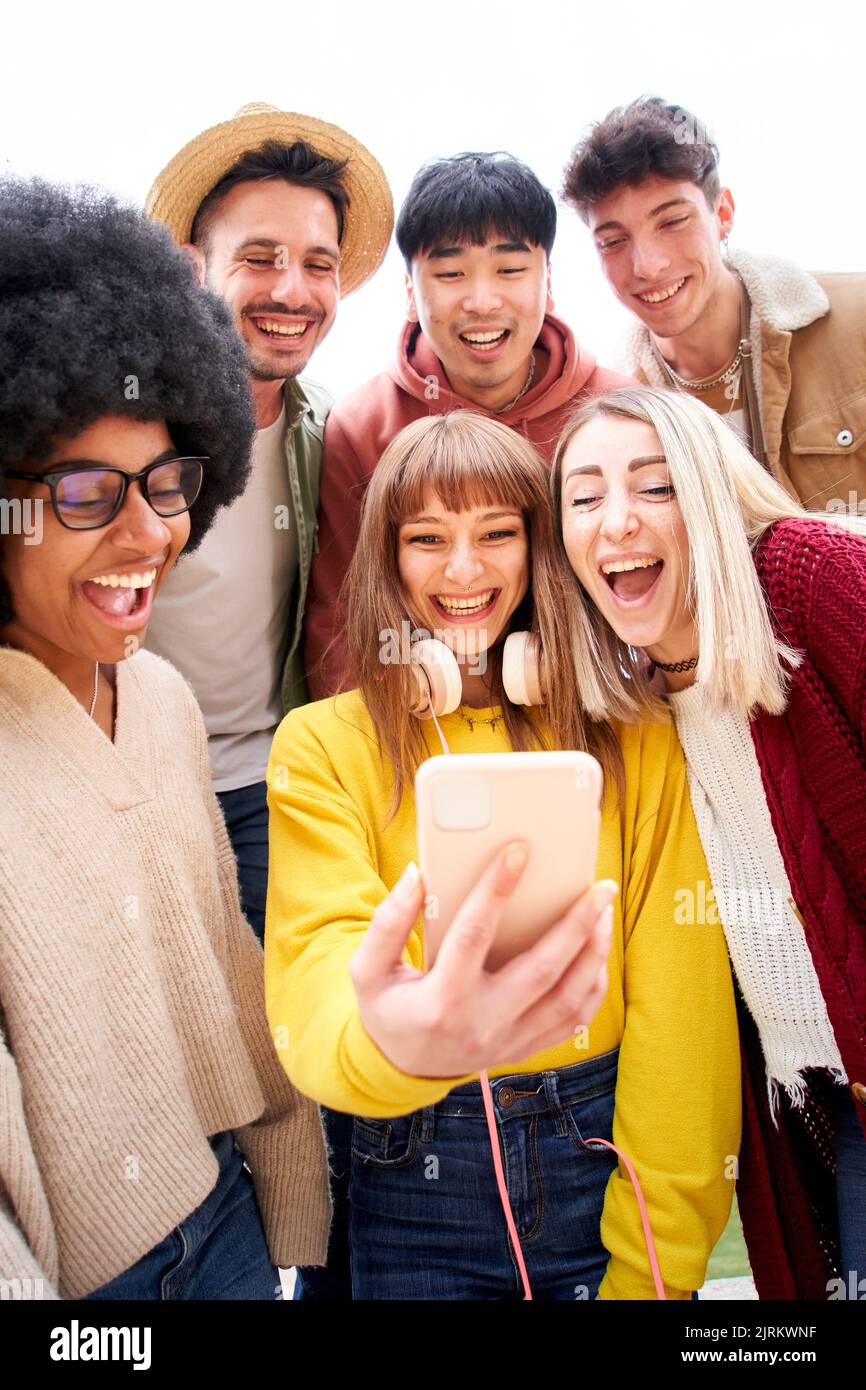 Vertikales Foto. Multikulturelle Gruppe von Freunden, die Handys benutzen und lachen - fröhliche Studenten Reihen sich an und schauen auf den Smartphone-Bildschirm. Jung Stockfoto