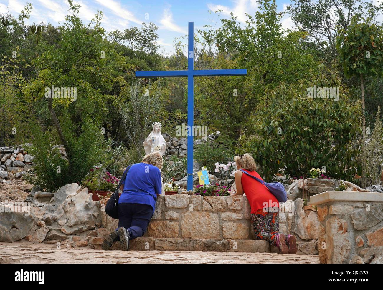 Zwei Frauen knien im Gebet am „Fuß“ des Podbrdo (Erscheinungsberg), vor dem blauen Kreuz und der Statue der seligen Jungfrau Maria (Medjugorje) Stockfoto