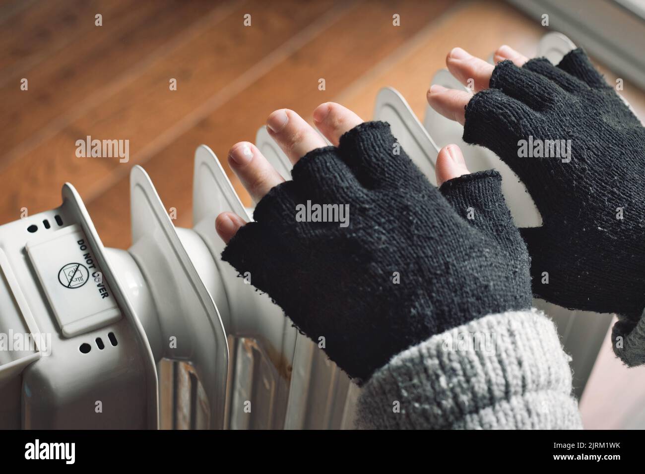 Nahaufnahme der Hände einer Person, die eine Wolljacke und schwarze wollige fingerlose Handschuhe in der Nähe eines Heizkörpers trägt, der versucht, sich im Winter warm zu halten Stockfoto