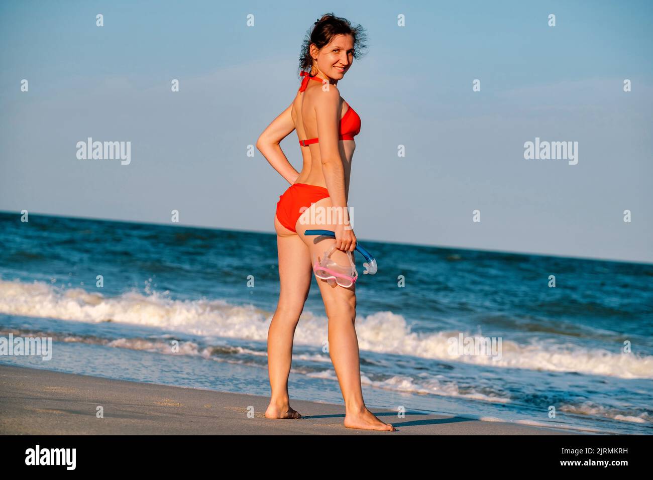 Sandgebräunte Frau mit Schnorchelausrüstung im Hintergrund des Strandes. Glücklicher Lebensstil. Fröhliche Frau im Bikini, die am Strand bis zum Meer steht. Blu Stockfoto