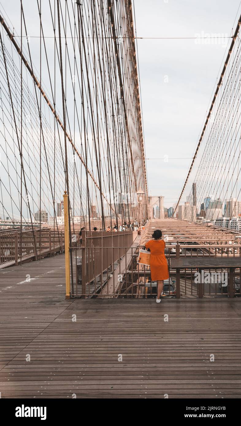 Eine vertikale Aufnahme einer Frau in einem orangefarbenen Kleid auf der Brooklyn Bridge, New York Stockfoto