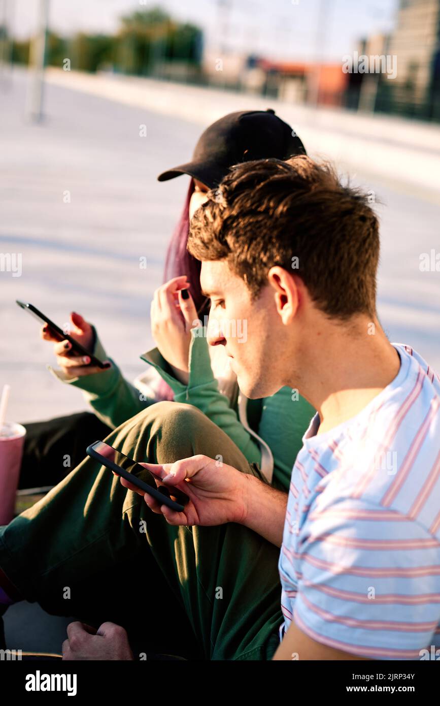 Zwei Jugendliche, die auf Social Media-Seiten sitzen und Telefone benutzen, sitzen im städtischen Außenbereich. Stockfoto