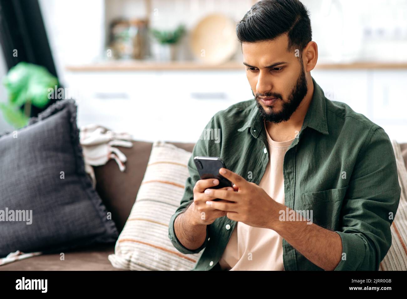 Verwirrten indischen oder arabischen Kerl in Freizeitkleidung, sitzt auf dem Sofa in einem Wohnzimmer, mit Smartphone, SMS online, in sozialen Netzwerken surfen, Nachrichten lesen, Sieht den Bildschirm verwirrt an. Wireless-Technologien Stockfoto