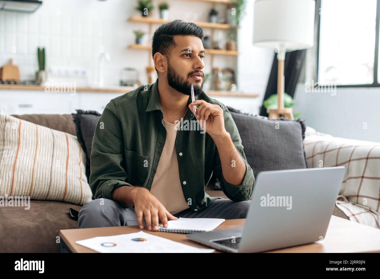 Nachdenklicher, kluger inder oder araber, Student oder Freiberufler, mit Laptop, ferngesteuert arbeiten oder studieren, Webinar anhören, Notizen machen, Hausaufgaben machen, zu Hause auf dem Sofa sitzen, wegschauen, denken Stockfoto