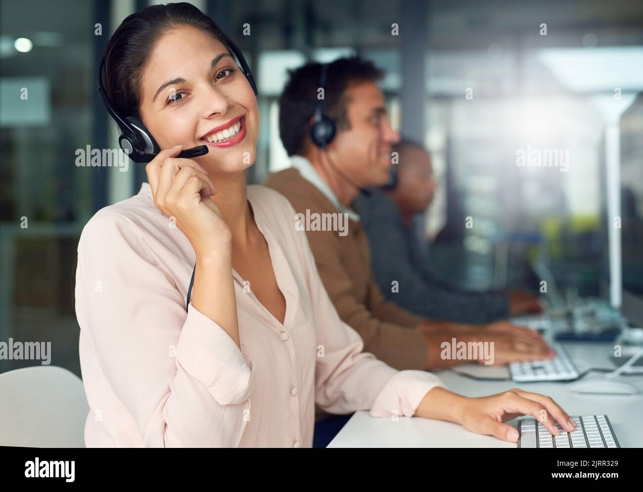 Rufen Sie uns jetzt an, um Rat von Experten zu erhalten. Porträt einer Callcenter-Agenten, die in einem Büro mit ihren Kollegen im Hintergrund arbeitet. Stockfoto