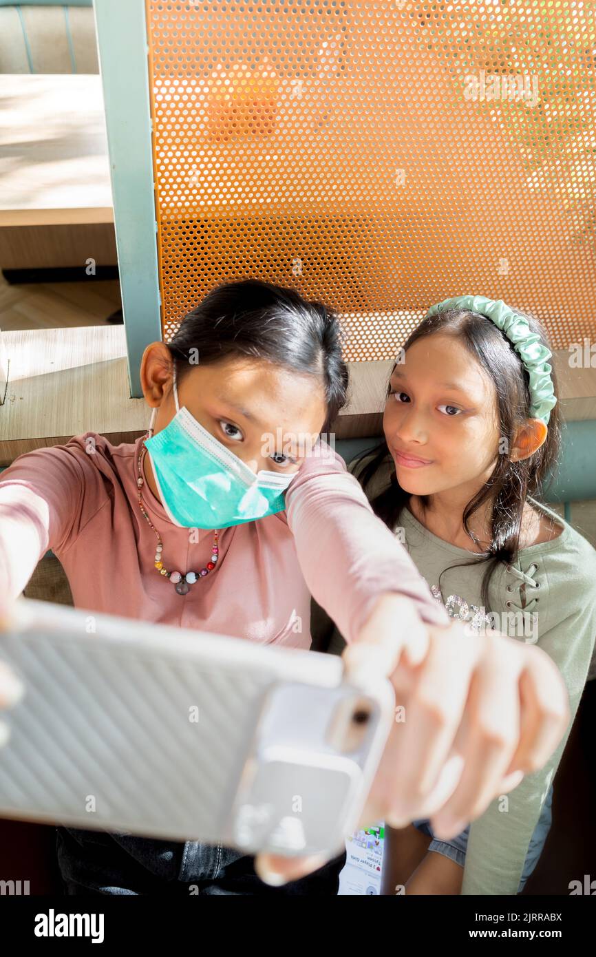 Asiatische Teenager-Mädchen tragen Gesichtsmaske und machen Selfie zusammen mit Schwester mit Smartphone in einem Restaurant Stockfoto