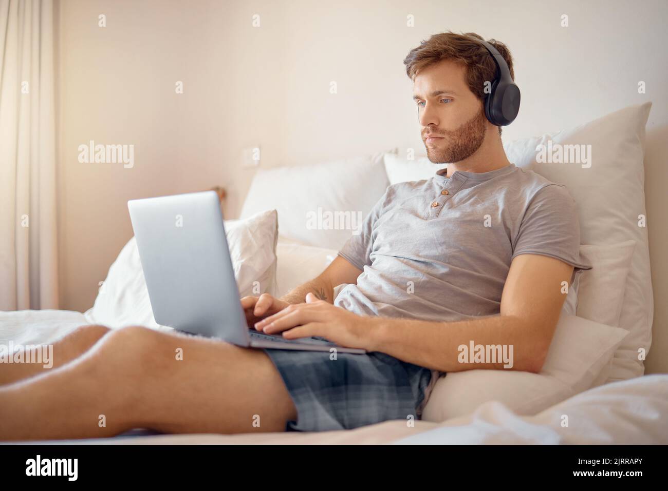 Mann, der am Laptop arbeitet oder schreibt und Musik mit Kopfhörern im Bett zu Hause hört. Telearbeiter streamen Podcast online, während sie lesen und senden Stockfoto