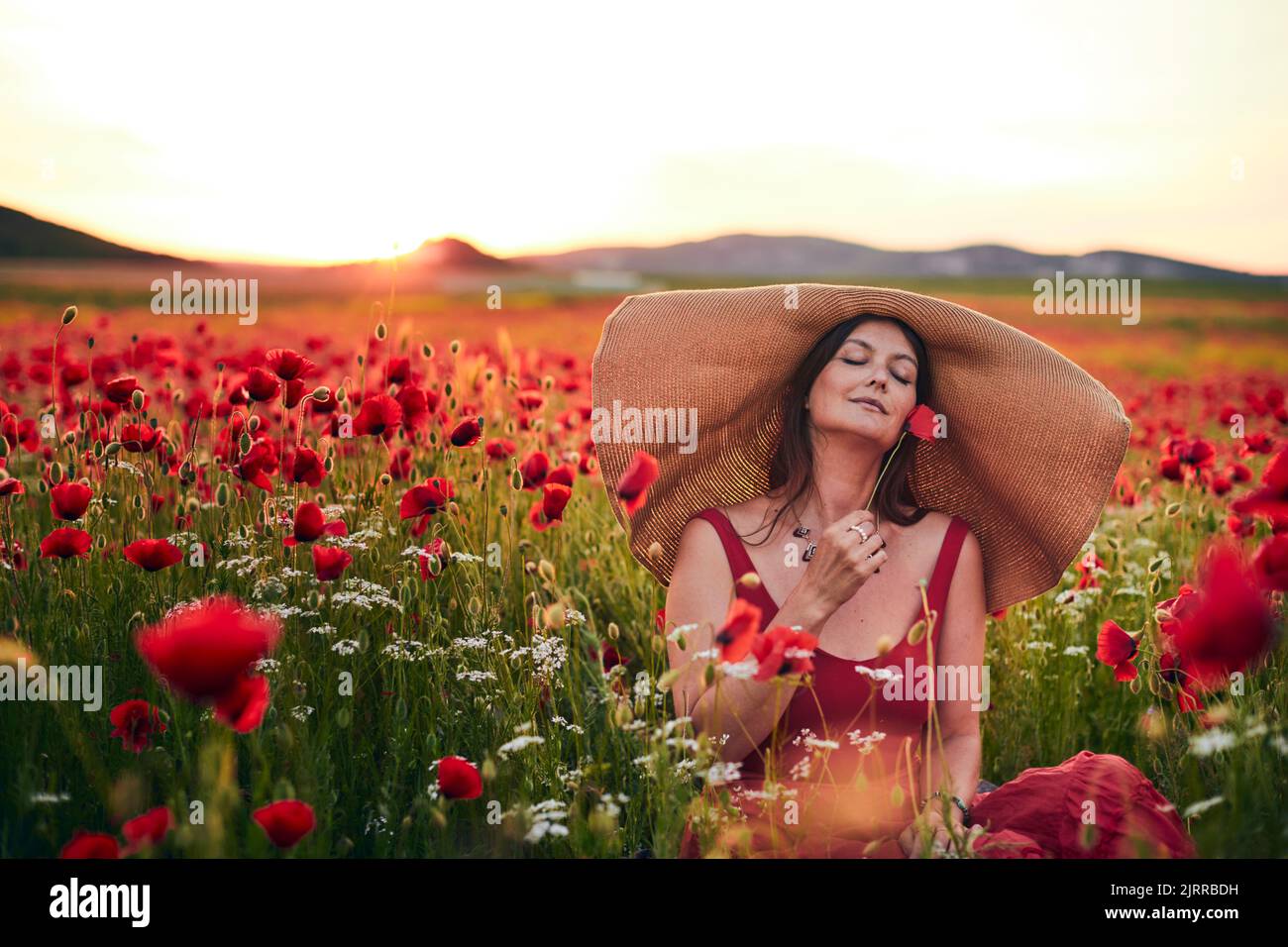 Porträt einer Frau in einem Mohnfeld, die bei Sonnenuntergang einen Hut mit breiter Krempe trägt. Vorderansicht. Stockfoto