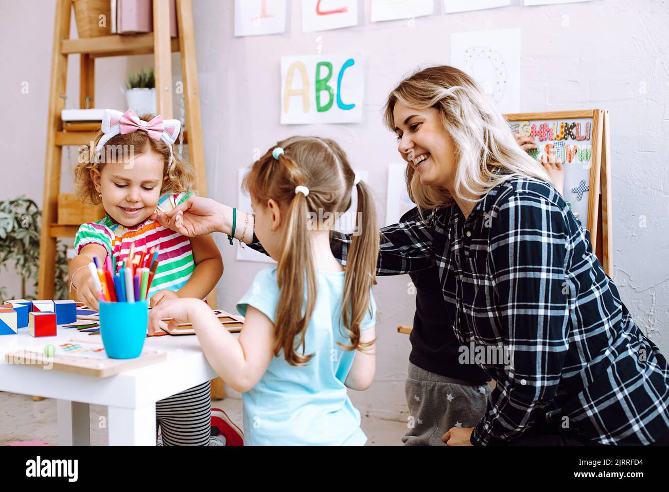 Porträt einer jungen lächelnden Frau, die am Tisch um spielende Kinder sitzt. Lehrer streckt Hand mit Papierschmetterling. Stockfoto