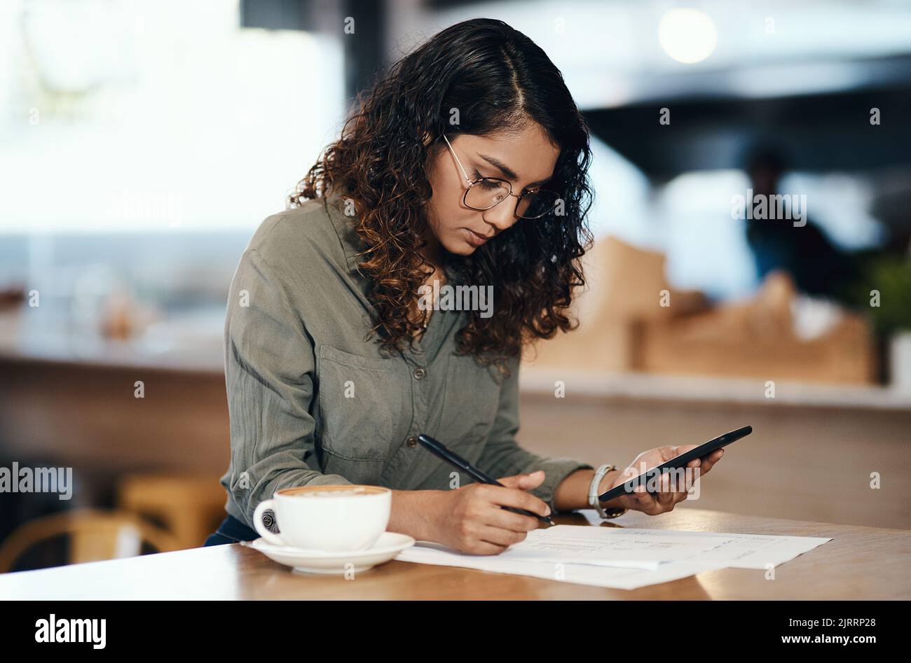 Inhaber oder Unternehmer eines Kleinunternehmens, der mit einem Telefon in der Hand Papier oder ein Dokument in einem Internetcafe oder Café ausfüllt. Junge Frau schreibt Stockfoto