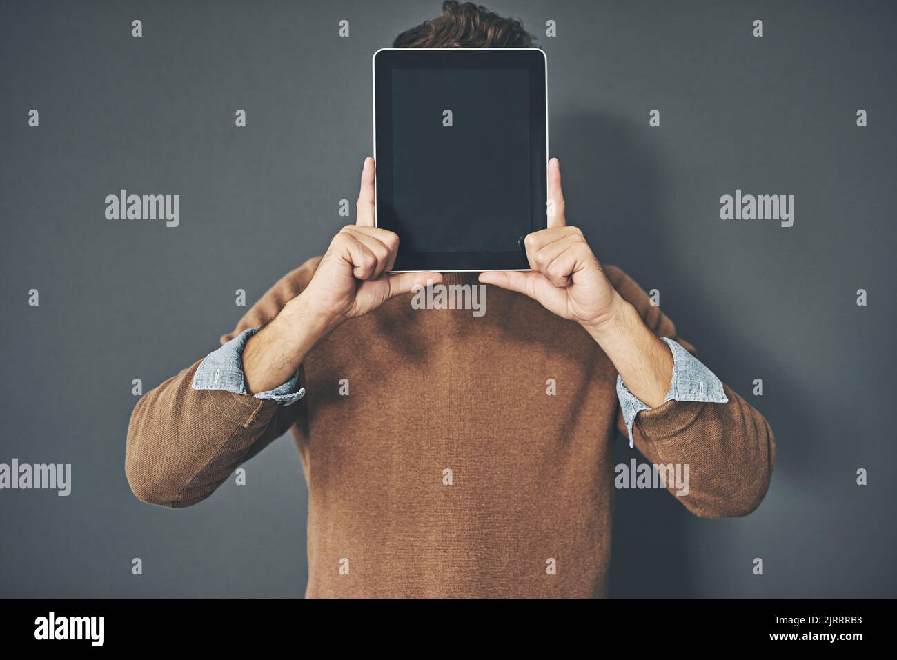 Junger Mann, der ein leeres Tablet vor sein Gesicht hält, in legerer Kleidung vor einem grauen Hintergrund mit Kopierraum. Alleinstehender Mann mit leerem Bildschirm Stockfoto