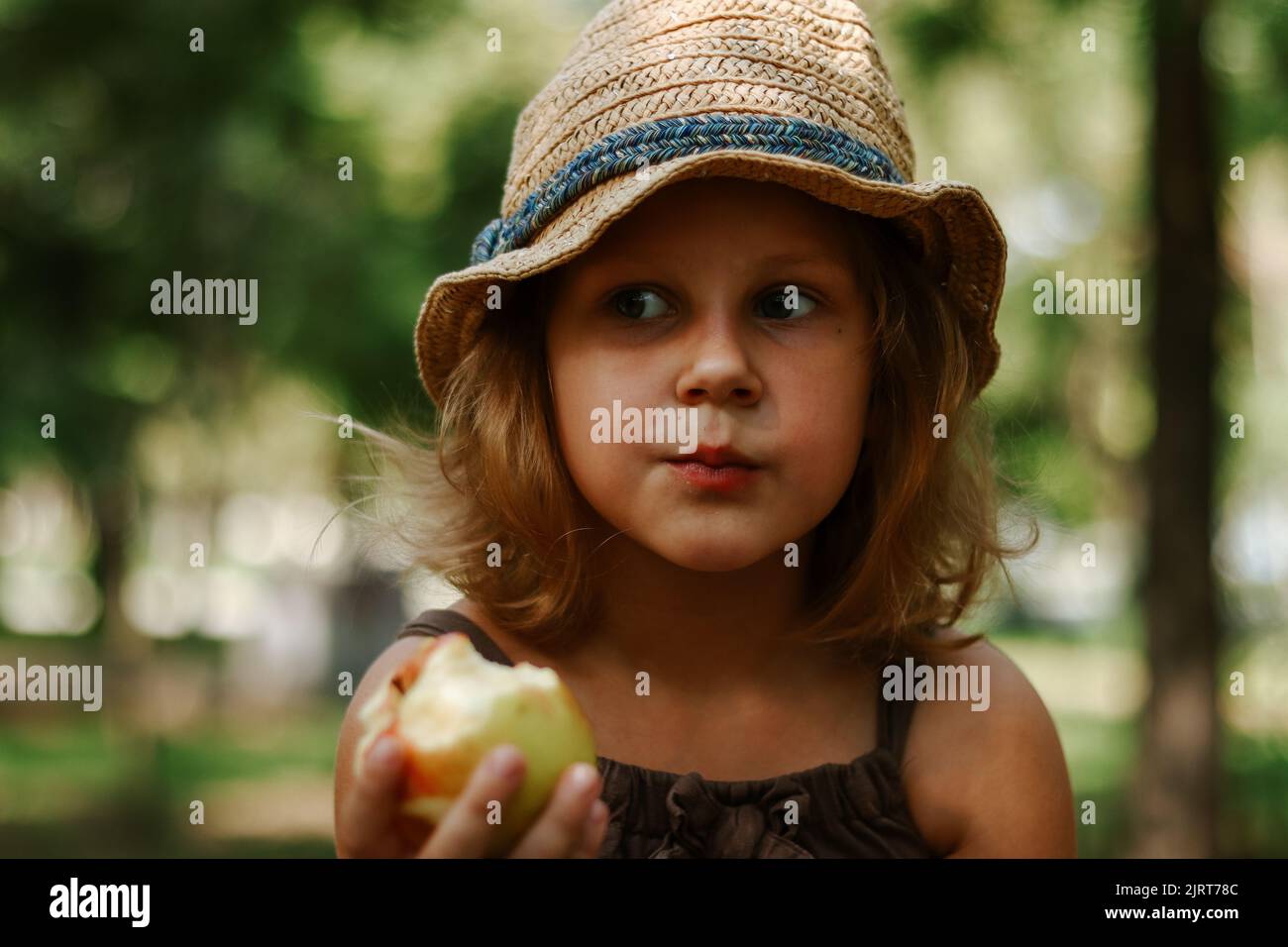 Porträt eines Kindes mit einem Apfel in den Händen. Baby 5 Jahre alt isst einen Apfel. Mädchen in einem Strohhut. Stockfoto