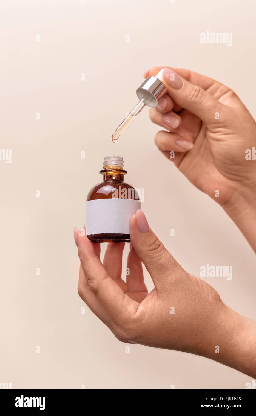 Hände halten Pipette und geöffnete Tropfflasche mit leerem Etikett über hellbeige, Nahaufnahme, Mockup. Naturkosmetik, Serum oder Essenz. Jeden Tag woma Stockfoto