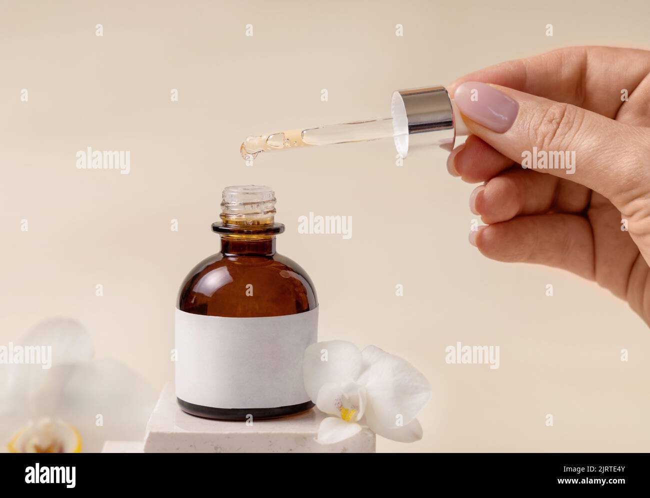Hand hält Pipette über geöffnete kosmetische Tropfflasche in der Nähe von weißen Orchideenblüten und Steinen auf hellbeige, Nahaufnahme, Mockup. Natürliche Hautpflege produ Stockfoto