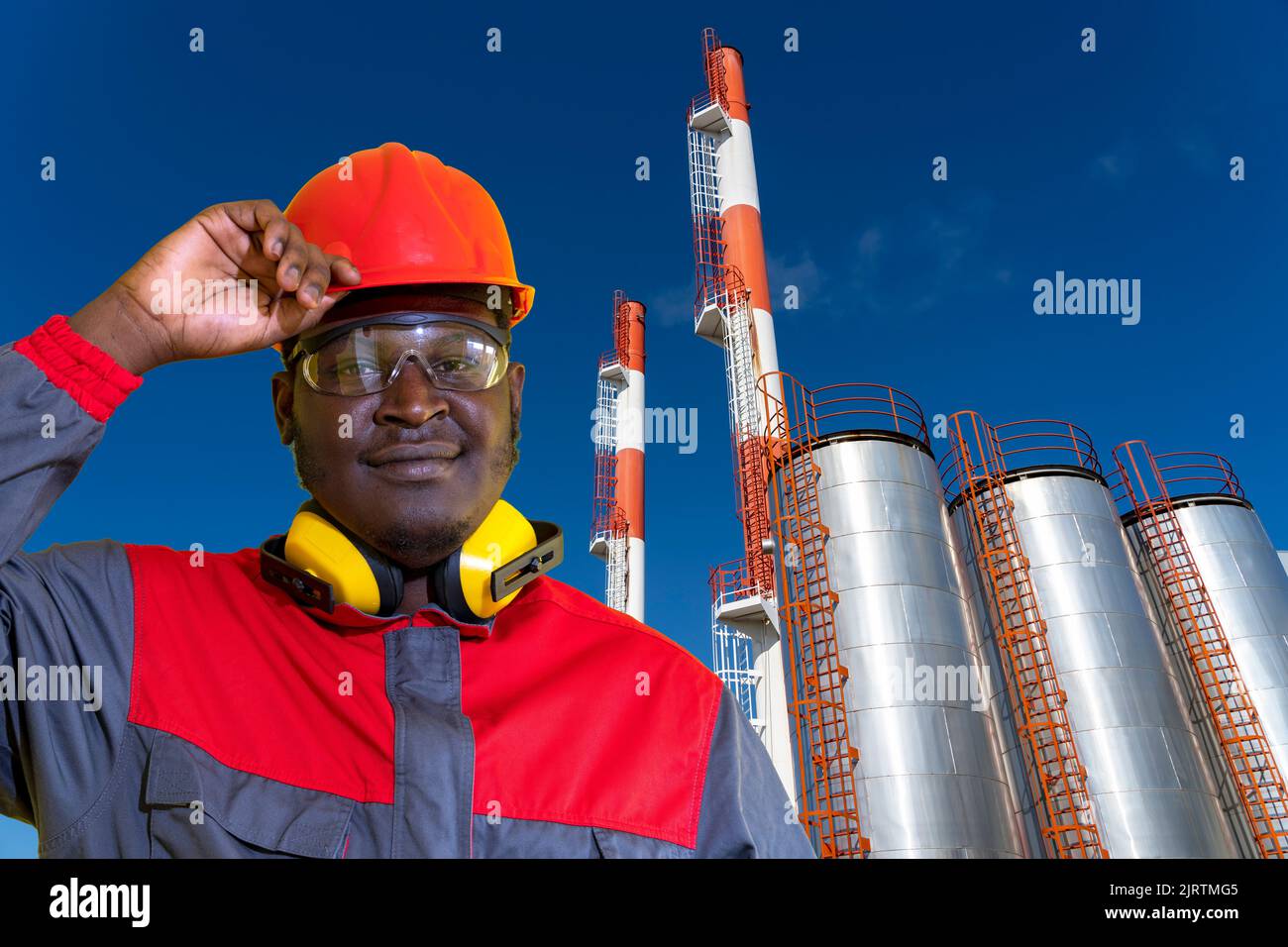 Junge Afroamerikanische Kraftwerks- Oder Ölarbeiter In Red Hardhat Stehen Vor Lagertanks Der Ölindustrie. Stockfoto
