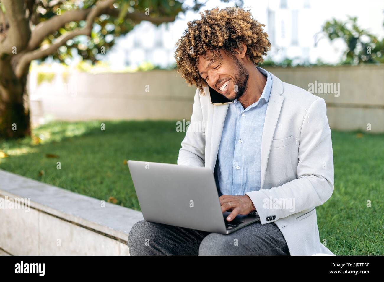 Positiv beschäftigt erfolgreicher hispanischer oder brasilianischer Geschäftsmann oder Top-Manager, arbeitet im Freien mit Laptop, spricht auf dem Smartphone mit dem Kunden oder Kollegen, tippt auf der Tastatur. Telefongespräch während der Arbeit Stockfoto